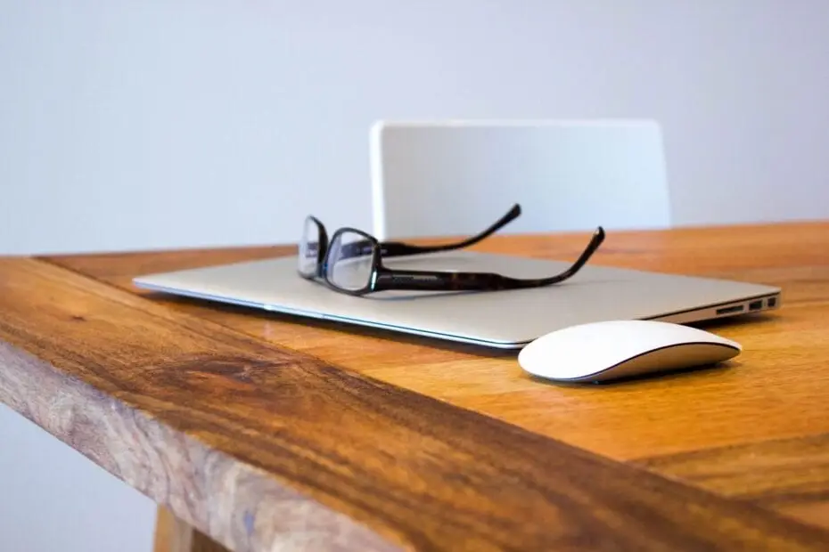 glasses on top of a closed laptop on top of a wooden desk with a white plastic chair behind